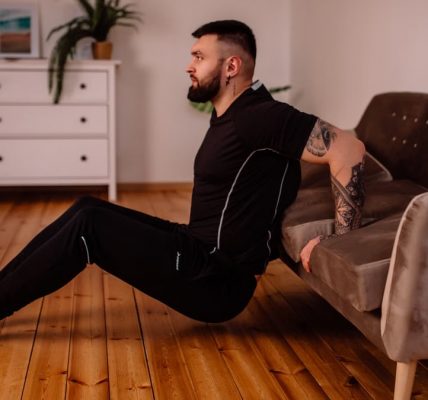 A bearded man in black fitness attire exercising at home, showcasing arm tattoos and a healthy lifestyle.