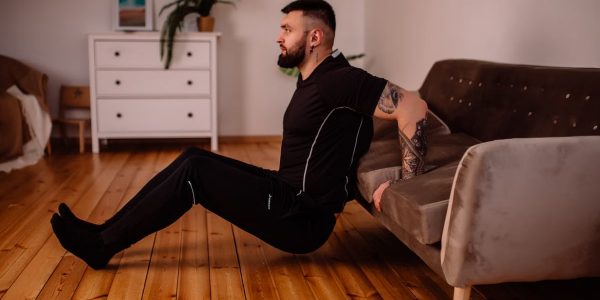 A bearded man in black fitness attire exercising at home, showcasing arm tattoos and a healthy lifestyle.